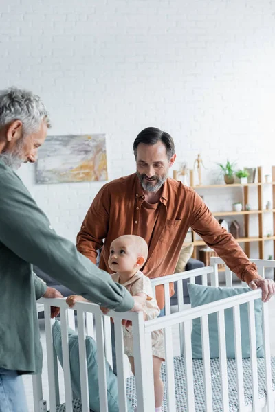 Smiling homosexual parents standing near baby daughter in crib — Stock Photo