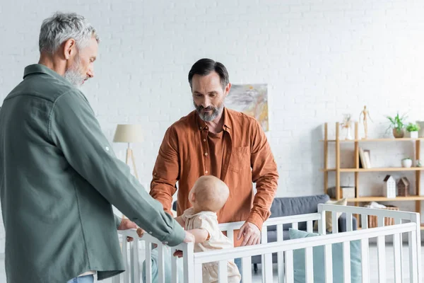 Homosexual fathers looking at baby in crib at home — Stock Photo