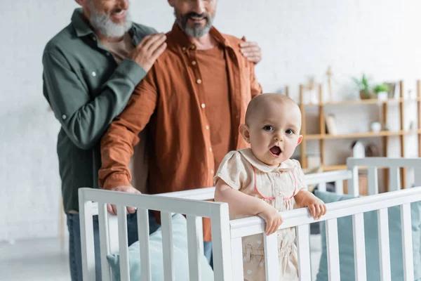 Toddler girl in baby bed near blurred homosexual parents — Stock Photo