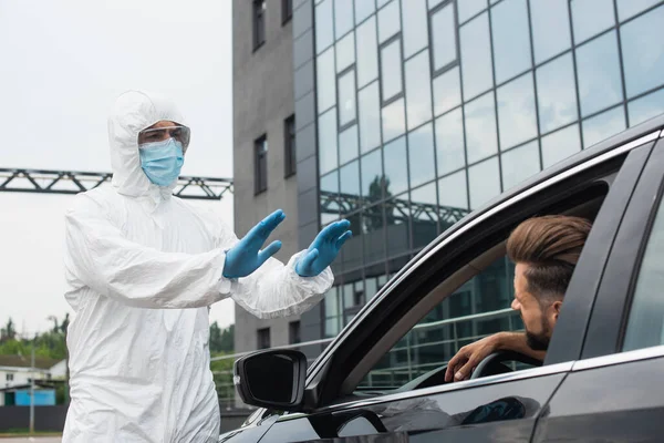 Trabajador médico en traje de materiales peligrosos que muestra gestos de parada al conductor en el coche - foto de stock