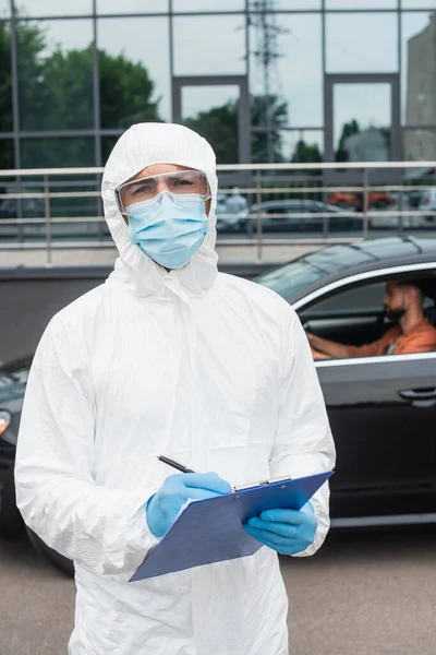 Border guard in hazmat suit holding clipboard near man in car outdoors — Stock Photo