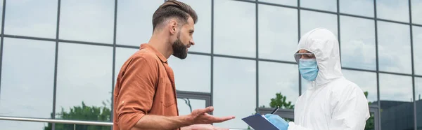 Mann im Gespräch mit medizinischem Personal im Schutzanzug, Schriftzug auf Klemmbrett im Freien, Banner — Stockfoto