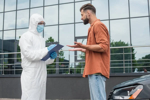 Hombre hablando con el guardia fronterizo en traje de materiales peligrosos escribiendo en el portapapeles cerca del coche - foto de stock