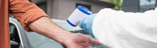 Обрізаний вид медичного працівника, який тримає пірометр біля людини і розмитий автомобіль, банер — стокове фото