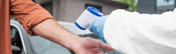 Ausgeschnittene Ansicht von medizinischem Personal mit Pyrometer in der Nähe von Menschen und verschwommenem Auto, Banner — Stockfoto