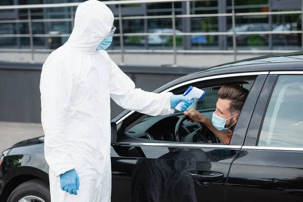 Trabajador médico en traje protector sosteniendo pirómetro cerca de hombre en máscara en coche - foto de stock