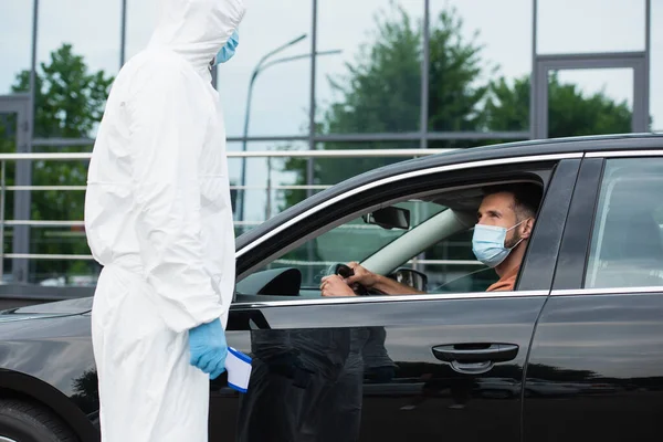 Medical worker in protective suit holding pyrometer near driver in car — Stock Photo