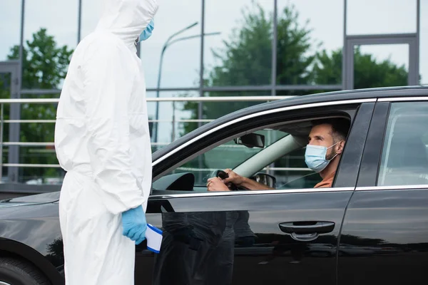 Travailleur médical en combinaison de protection tenant pyromètre près du conducteur dans la voiture — Photo de stock