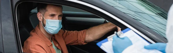 Водій у масці, дивлячись на розмитого медичного працівника, що пише на кишені, банер — стокове фото