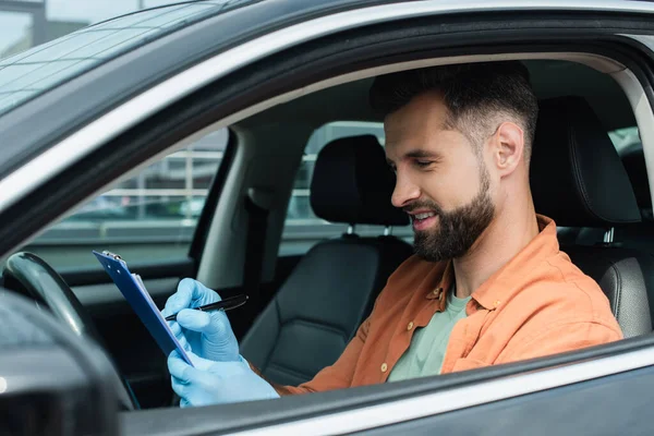 Motorista sorridente em luvas de látex escrevendo na área de transferência no carro — Fotografia de Stock