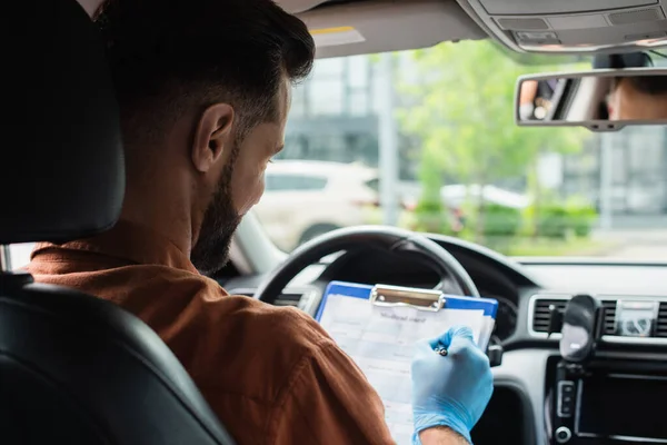 Driver in latex glove writing on clipboard in auto on blurred background — Stock Photo