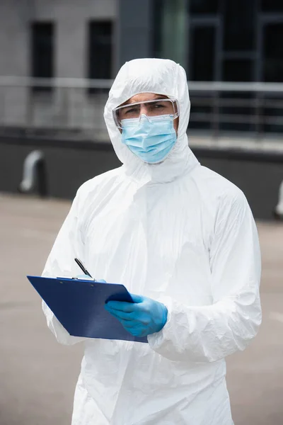 Trabalhador médico em terno hazmat e luvas de látex escrevendo na área de transferência e olhando para a câmera ao ar livre — Fotografia de Stock