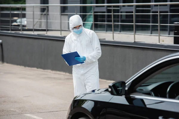 Trabalhador médico em terno hazmat e óculos escrevendo na área de transferência perto do carro embaçado — Fotografia de Stock