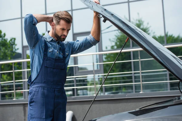 Nachdenklicher Mechaniker betrachtet Auto mit offener Motorhaube im Freien — Stockfoto