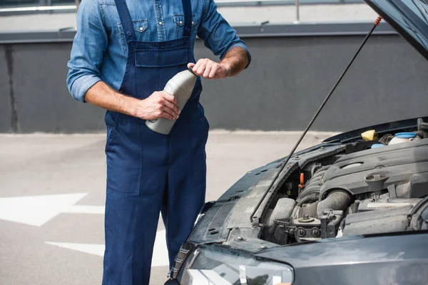 Cropped view of mechanic holding bottle of motor oil near car with open hood — Stock Photo