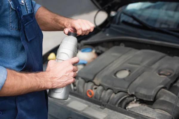 Vista recortada del mecánico en la botella de retención uniforme de aceite de motor cerca de coche borroso - foto de stock