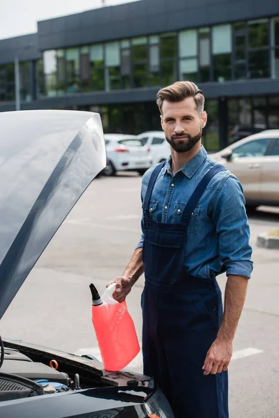 Mechanic with canister of windshield washer fluid looking at camera near auto with open hood — Stock Photo