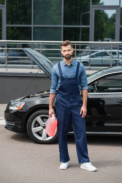 Riparatore in tuta che tiene il liquido della rondella del parabrezza vicino all'auto all'aperto — Foto stock