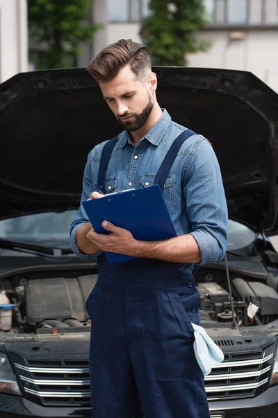 Mechanic writing on clipboard near blurred car outdoors — Stock Photo