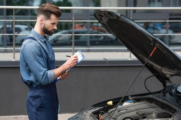 Vue latérale du mécanicien en salopette écrivant sur le presse-papiers près de la voiture à l'extérieur — Photo de stock