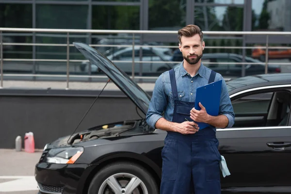 Mechaniker mit Klemmbrett und Kugelschreiber blickt auf Kamera in der Nähe von Auto im Freien — Stockfoto