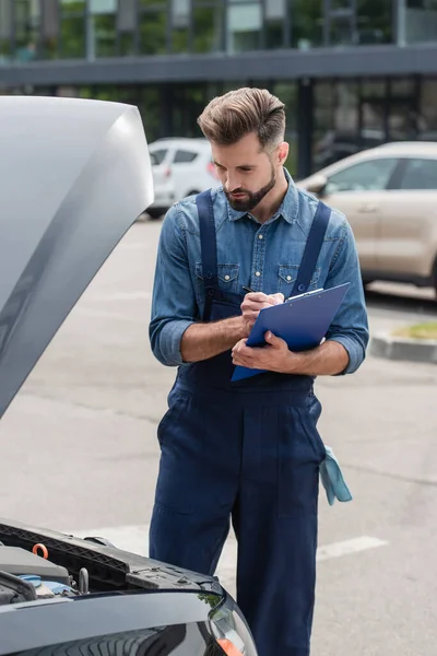 Mechanic writing on clipboard while looking at engine of car outdoors — Stock Photo