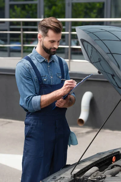 Mechanic in overalls writing on clipboard near car with open hood — Stock Photo