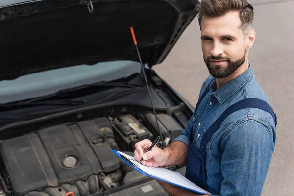 Hochwinkelaufnahme des Mechanikers, der auf Kamera schaut, während er auf Klemmbrett in der Nähe des Autos auf verschwommenem Hintergrund im Freien schreibt — Stockfoto