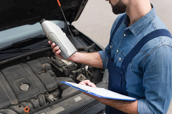 Vue recadrée du mécanicien tenant l'huile moteur et le presse-papiers près de la voiture — Photo de stock