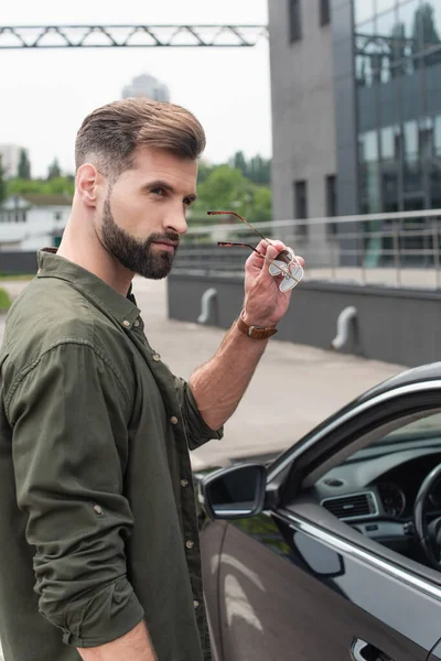 Giovane uomo che tiene gli occhiali da sole vicino auto offuscata all'aperto — Foto stock