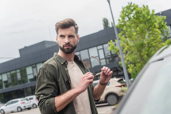 Uomo barbuto che tiene gli occhiali da sole vicino auto sfocata — Foto stock