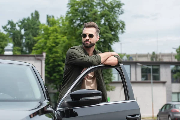 Homme barbu dans des lunettes de soleil debout près de la voiture avec porte ouverte — Photo de stock