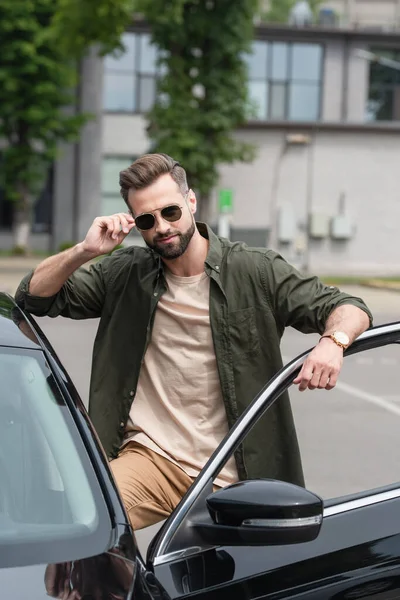 Bearded man adjusting sunglasses near auto outdoors — Stock Photo