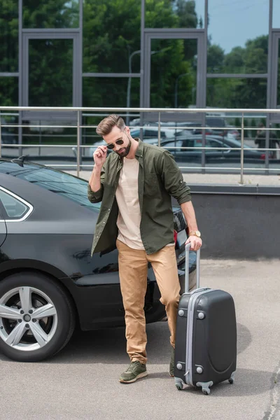 Tourist in sunglasses holding suitcase near car outdoors — Stock Photo