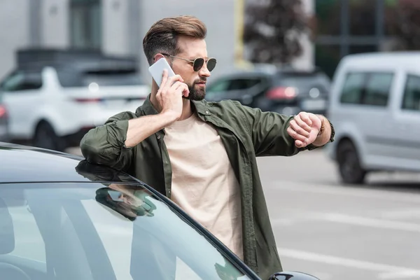 Man in sunglasses looking at wristwatch while talking on smartphone near car — Stock Photo