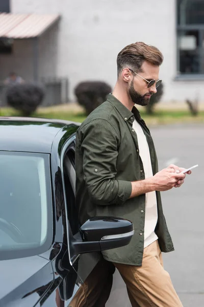 Side view of man in sunglasses using smartphone near car — Stock Photo