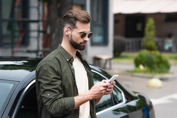 Homem de óculos de sol usando telefone celular perto do carro ao ar livre — Fotografia de Stock