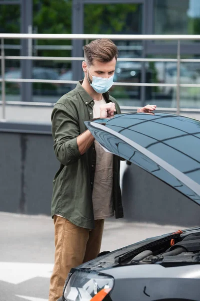 Driver in medical mask opening hood of car outdoors — Stock Photo