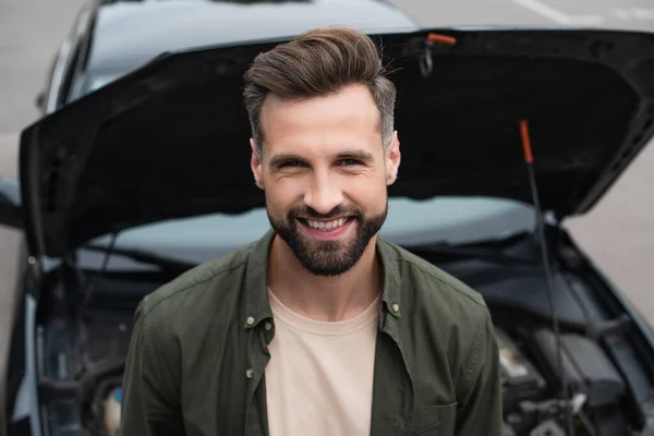Hombre positivo sonriendo a la cámara cerca del coche sobre fondo borroso - foto de stock