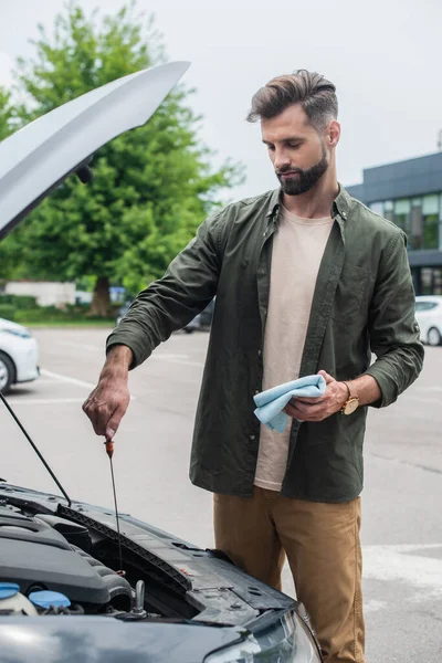 Uomo barbuto che controlla olio motore in auto e tiene straccio all'aperto — Foto stock