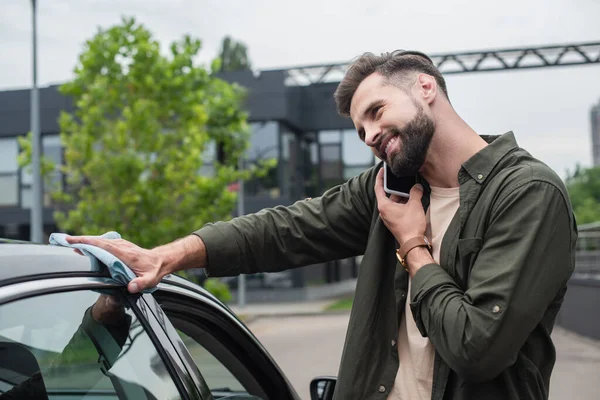 Smiling man talking on smartphone while wiping car outdoors — Stock Photo