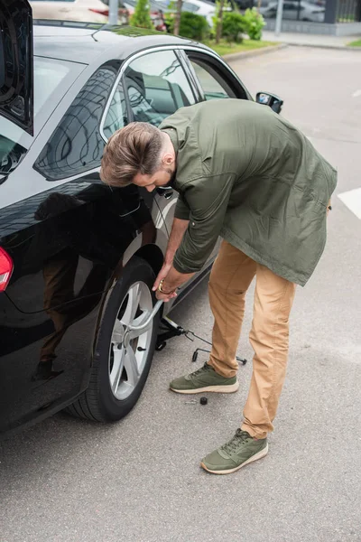 Young man using wheel wrench near lift jack under car — Stock Photo