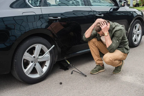 Displeased driver sitting near lift jack and wheel wrench on car — Stock Photo