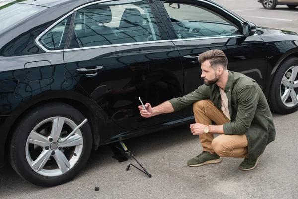 Vista lateral del hombre tomando una foto de la llave de la rueda cerca del coche con gato elevador al aire libre - foto de stock