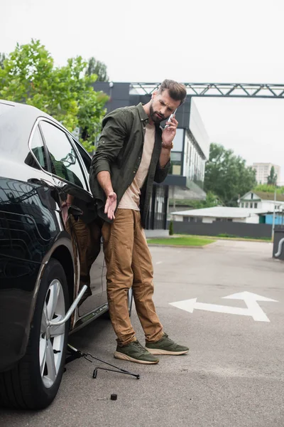 Man talking on smartphone near lift jack and wheel wrench in car — Stock Photo