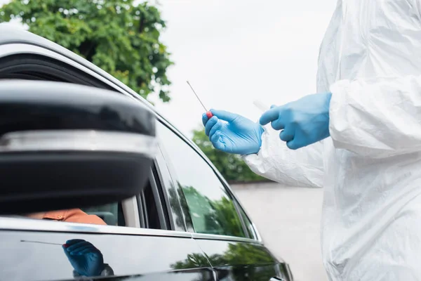 Vue recadrée du travailleur médical en combinaison de danger tenant test pcr près du conducteur dans une voiture floue — Photo de stock