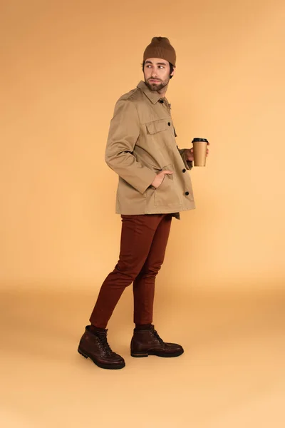 Full length view of man in stylish autumn clothes walking with takeaway drink on beige — Stock Photo