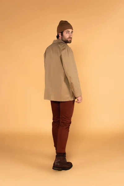 Full length view of fashionable man in beanie, jacket and brown pants looking at camera on beige — Fotografia de Stock