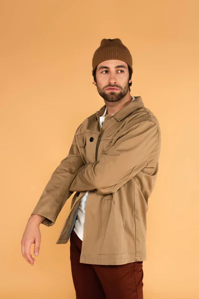 Молодой человек в куртке и коричневой шапочке смотрит в сторону, стоя изолированным на бежевом — стоковое фото