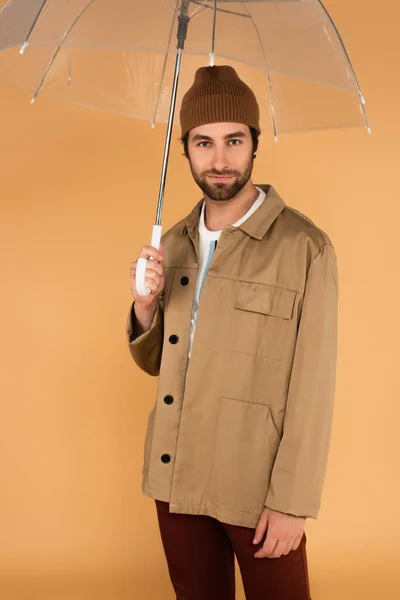 Мужчина в стильной осенней одежде смотрит в камеру под прозрачным зонтиком, изолированным от бежевого — стоковое фото